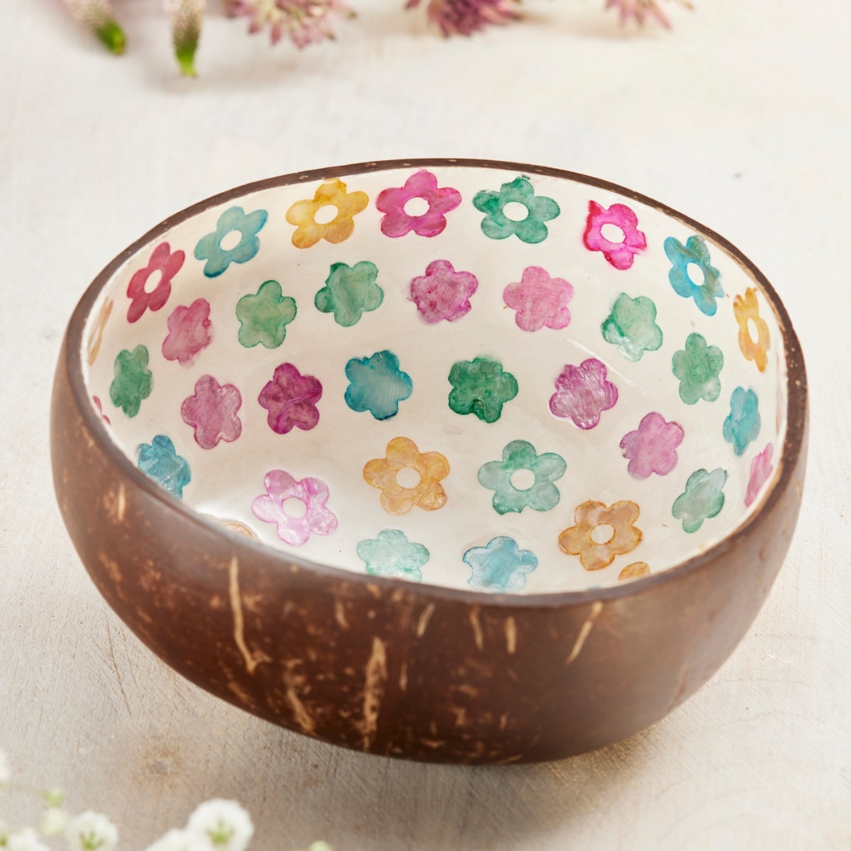 A decorative bowl made from a brown coconut shell, featuring a white interior decorated with a colorful pattern of hand-painted flowers in pink, green, blue, and yellow.