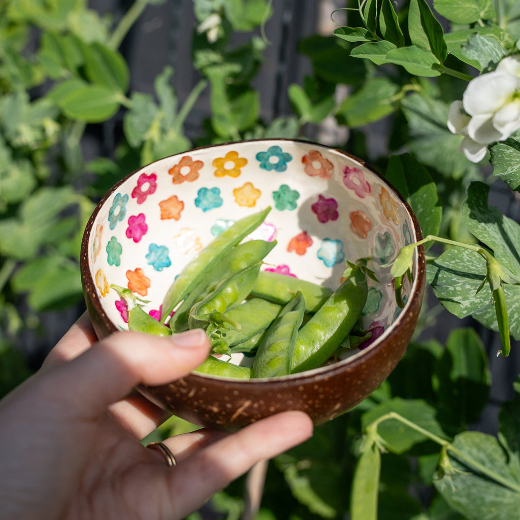 A hand holds a small speckled brown ceramic bowl filled with green pea pods, decorated with a colorful floral pattern inside, set against a background of green garden plants.