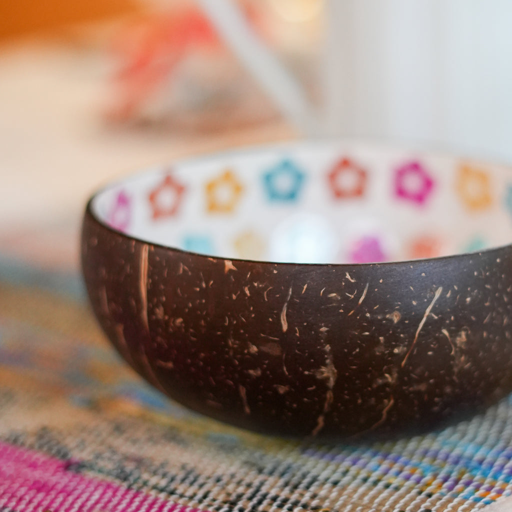 A coconut shell bowl with a dark brown textured exterior and a white interior decorated with colorful star patterns, resting on a woven multicolor placemat.