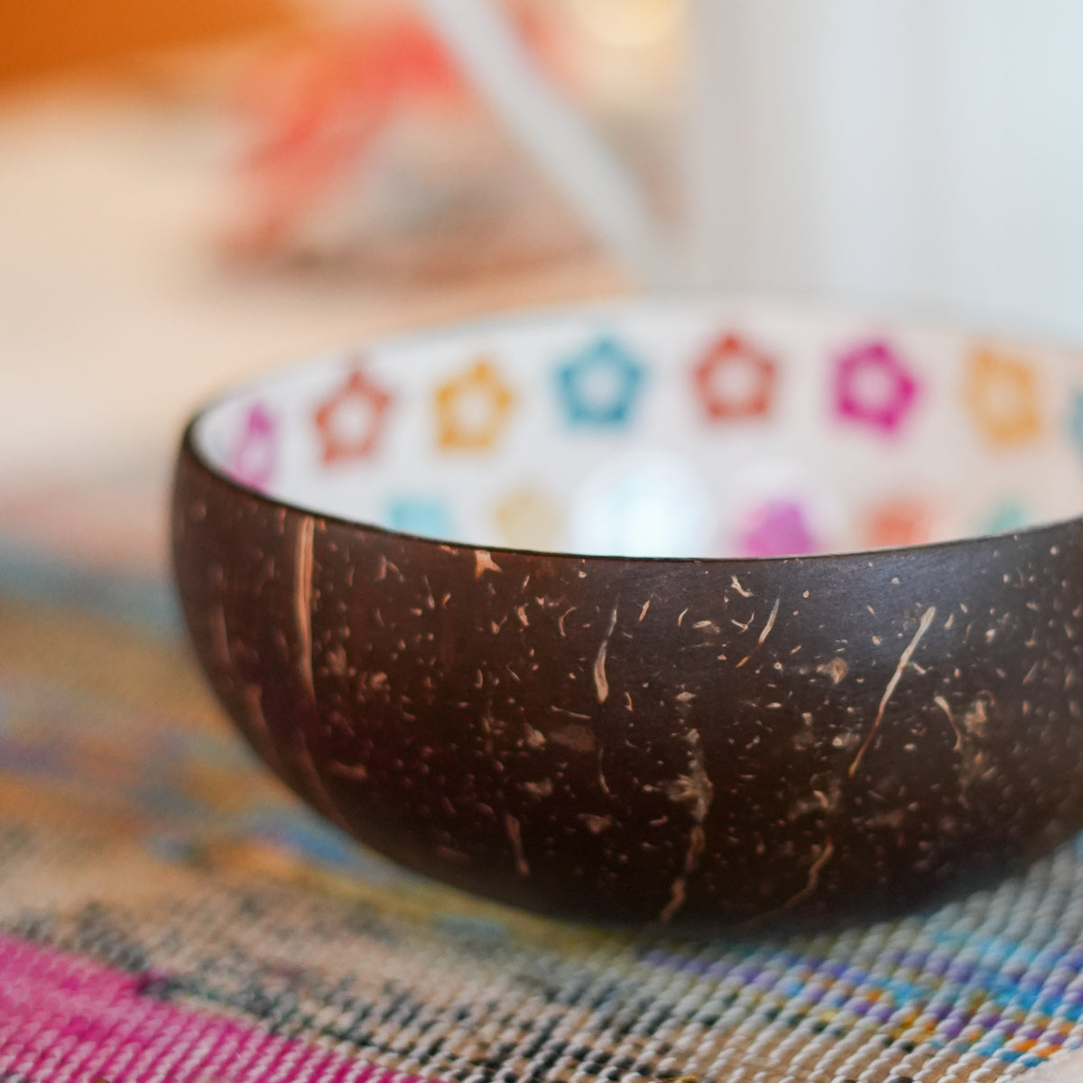 A coconut shell bowl with a dark brown textured exterior and a white interior decorated with colorful star patterns, resting on a woven multicolor placemat.