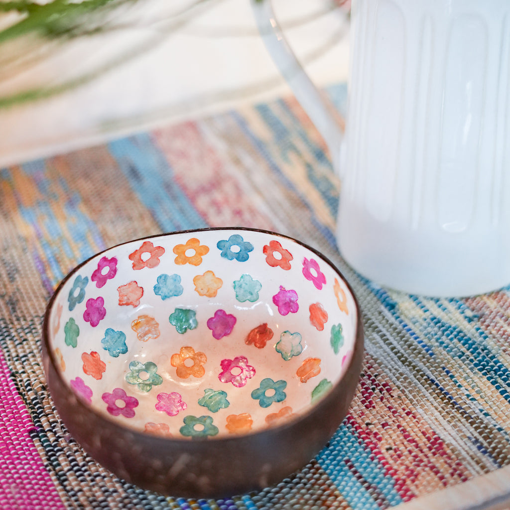 A small handmade bowl with a dark brown exterior and a white interior decorated with colorful painted flowers, set on a woven placemat next to a white pitcher.