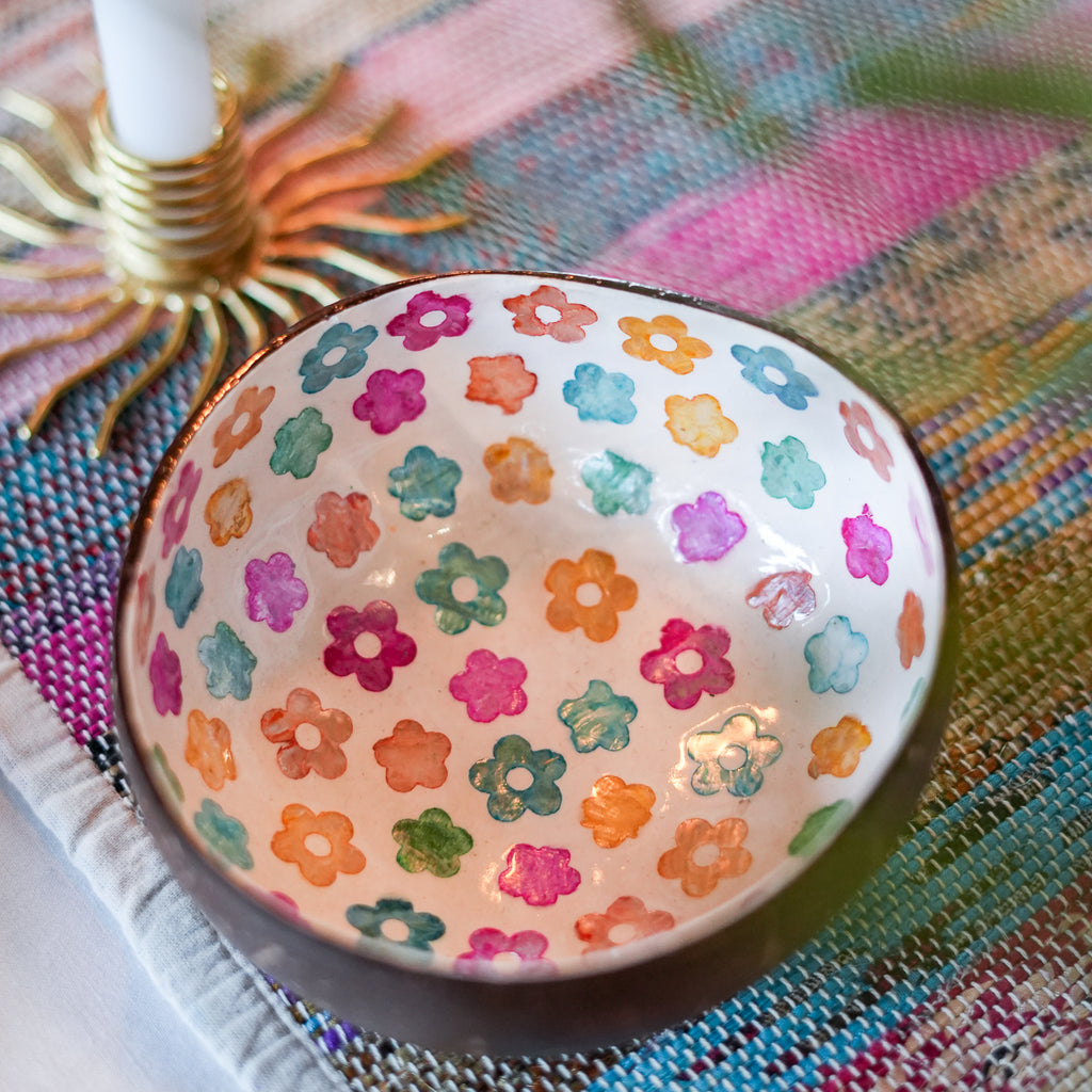A handmade ceramic bowl with a colorful hand-painted floral pattern inside, resting on a woven multicolor placemat next to a candle holder.
