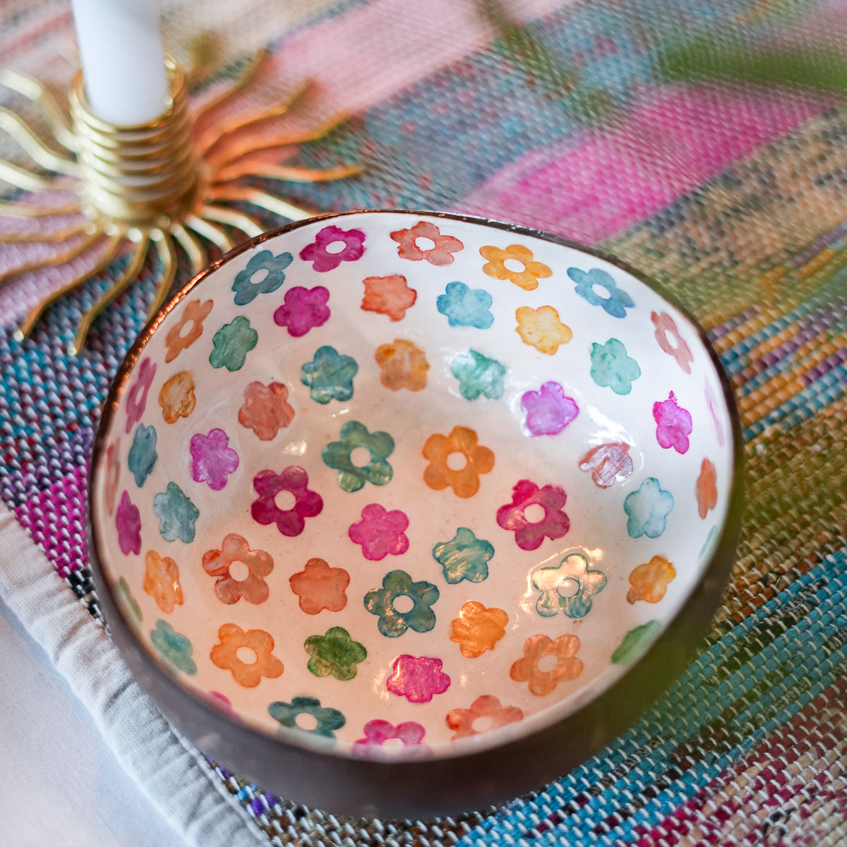 A handmade ceramic bowl with a colorful hand-painted floral pattern inside, resting on a woven multicolor placemat next to a candle holder.