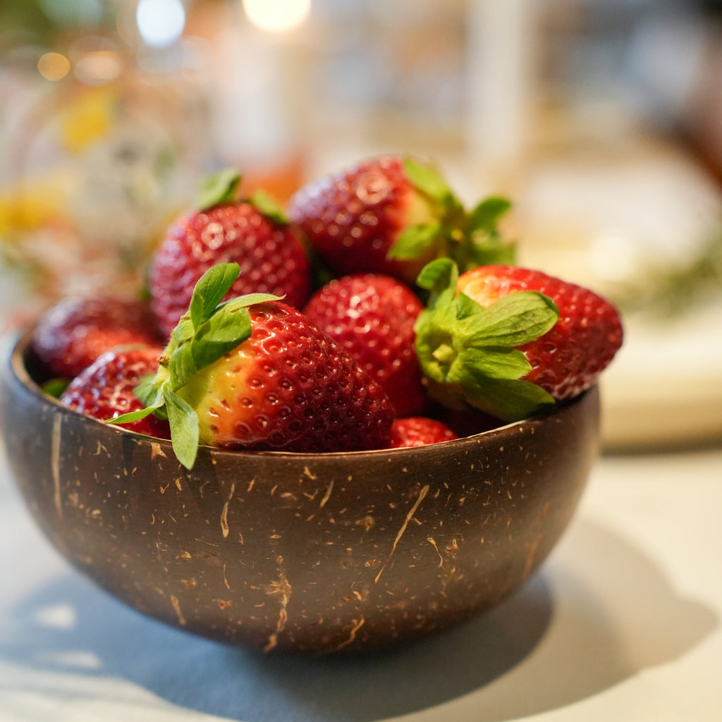 Fresh red strawberries with green tops piled in a dark brown coconut shell bowl on a light surface with a softly blurred background.