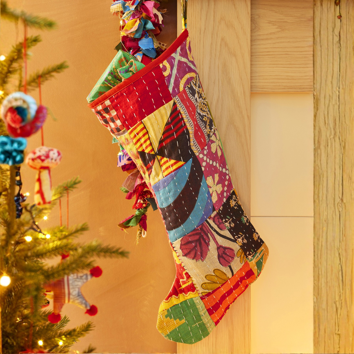 A colorful Christmas stocking made of patchwork quilted fabric hangs from a wooden door frame next to a decorated Christmas tree with warm string lights.