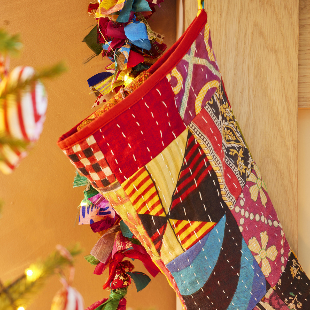 A colorful patchwork Christmas stocking hanging from a gold hook, decorated with vibrant fabric panels in red, yellow, blue, black, and pink, set against a warm wooden background.