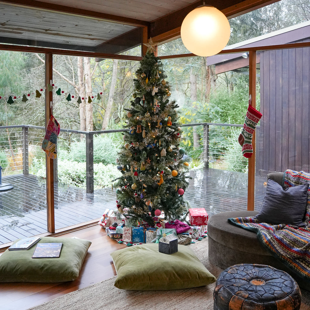 A decorated Christmas tree stands in a rustic indoor-outdoor living space with large windows overlooking a wooded deck. The tree is adorned with ornaments and wrapped gifts at the base, with pillows, blankets, and a pouf in the foreground.
