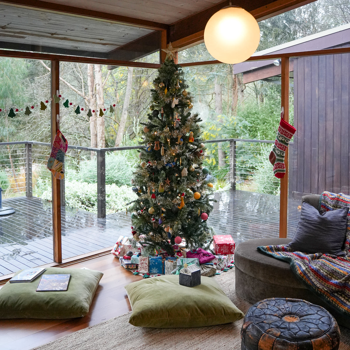 A decorated Christmas tree stands in a rustic indoor-outdoor living space with large windows overlooking a wooded deck. The tree is adorned with ornaments and wrapped gifts at the base, with pillows, blankets, and a pouf in the foreground.