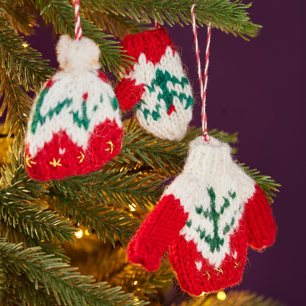 Three hand-knit Christmas mitten ornaments in red, white, and green hanging on a pine tree with warm string lights against a dark purple background.