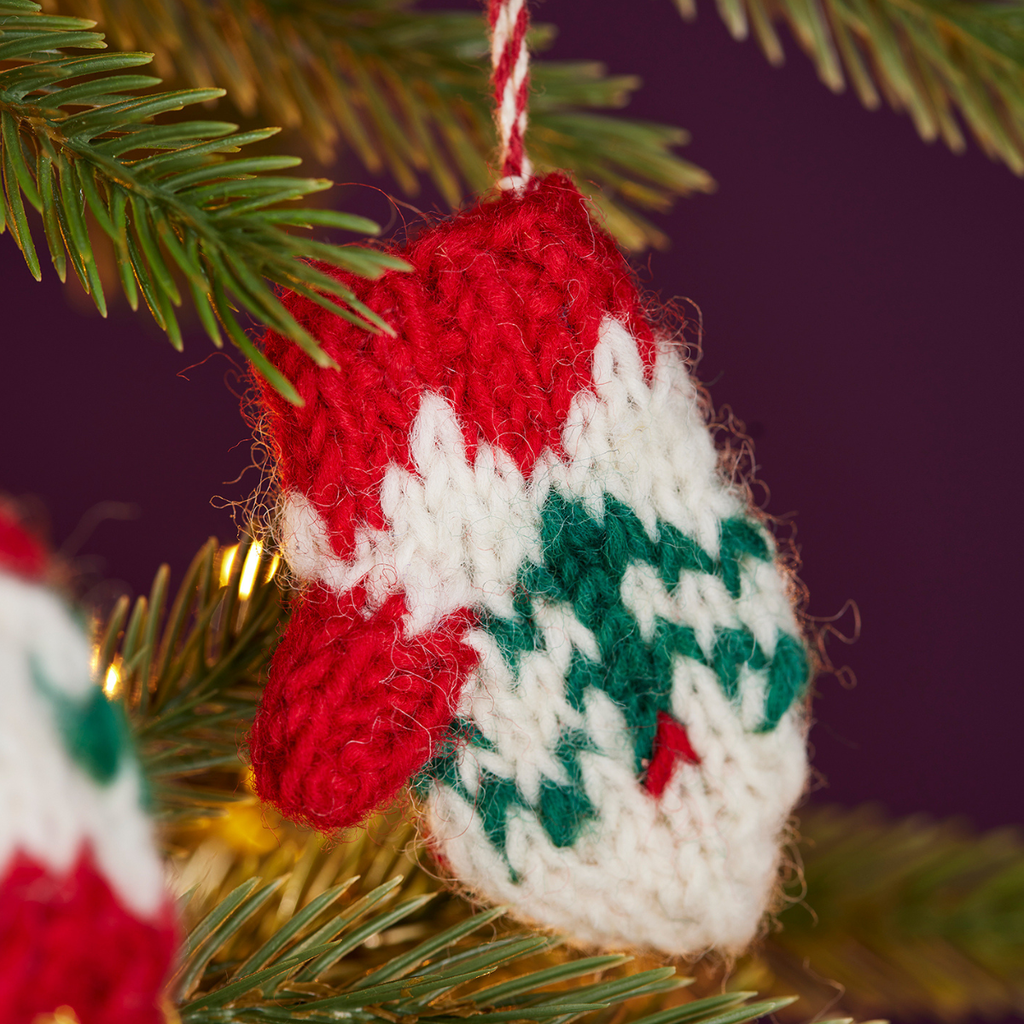 A close-up of a hand-knit Christmas ornament shaped like a mitten, hanging from a pine tree branch. The mitten ornament is red and white with a green chevron pattern on the lower half, and a small red nose detail.