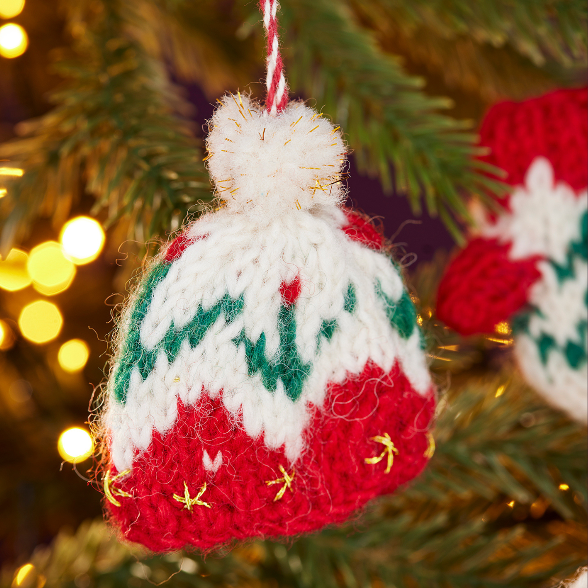 A close-up of a hand-knit Christmas ornament shaped like a winter hat, hanging on a Christmas tree. The ornament features a white top with a fluffy pom-pom, green zigzag patterns, and a red bottom with small gold embroidered details.