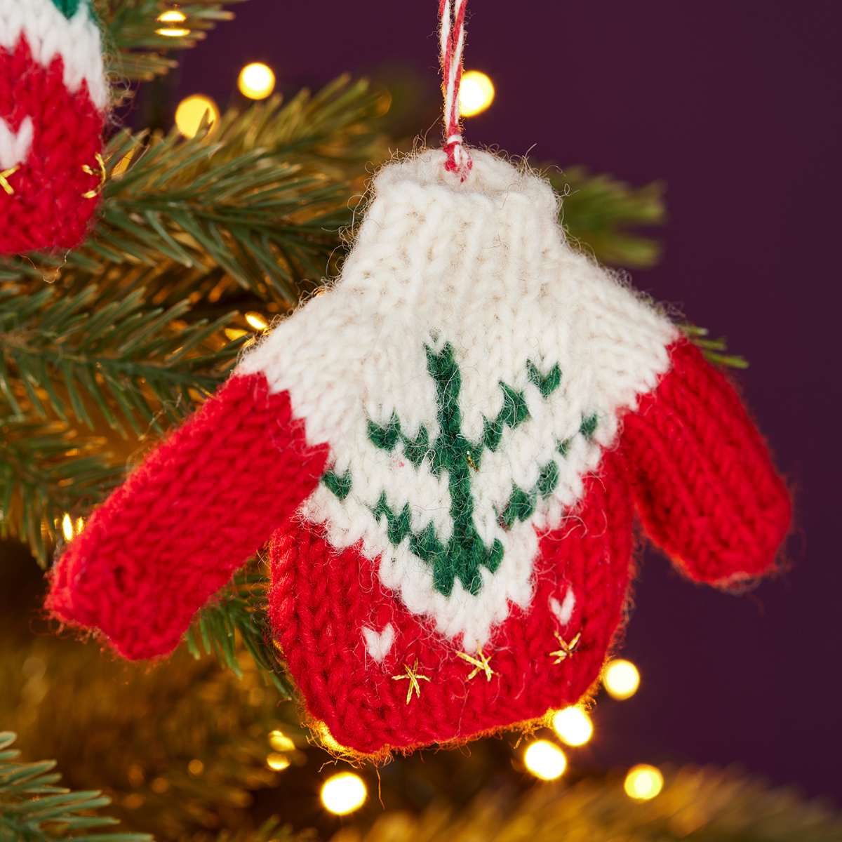A hand-knit Christmas ornament shaped like a mitten, featuring a white upper section with a green embroidered tree design, red sleeves and lower body, and small gold star accents. It is hanging on a Christmas tree branch with warm string lights in the background.