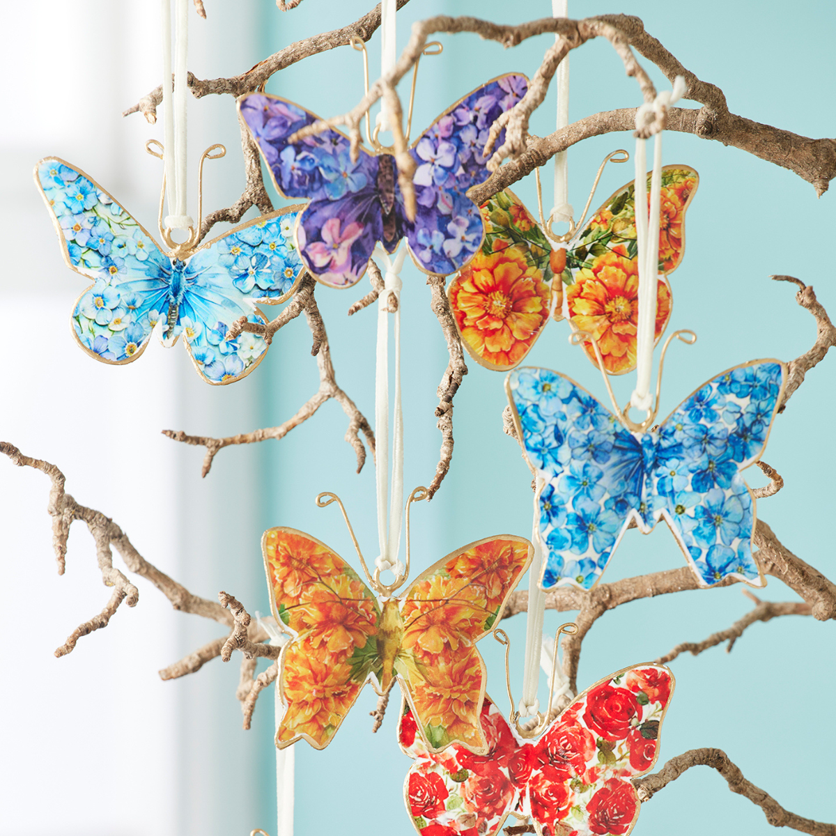 A set of decorative hanging ornaments shaped like butterflies, decorated with floral and patterned designs in blue, purple, orange, and red, displayed on a rustic branch against a light blue background.