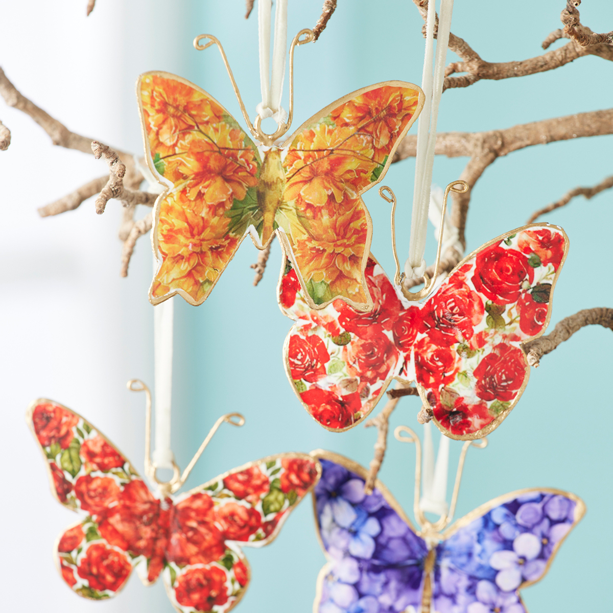 Three decorative butterfly ornaments with floral patterns hang from clear strings against a light blue background with thin brown branches.