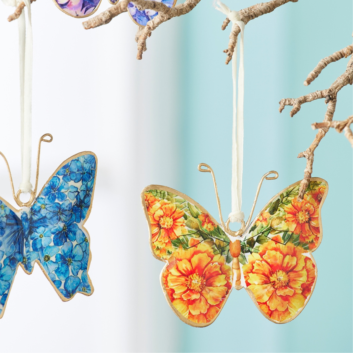 Decorative hanging butterfly ornaments displayed on a branch against a soft white and light blue background. One ornament on the left is blue with floral detailing, and the one on the right features orange and yellow flowers with green leaves.