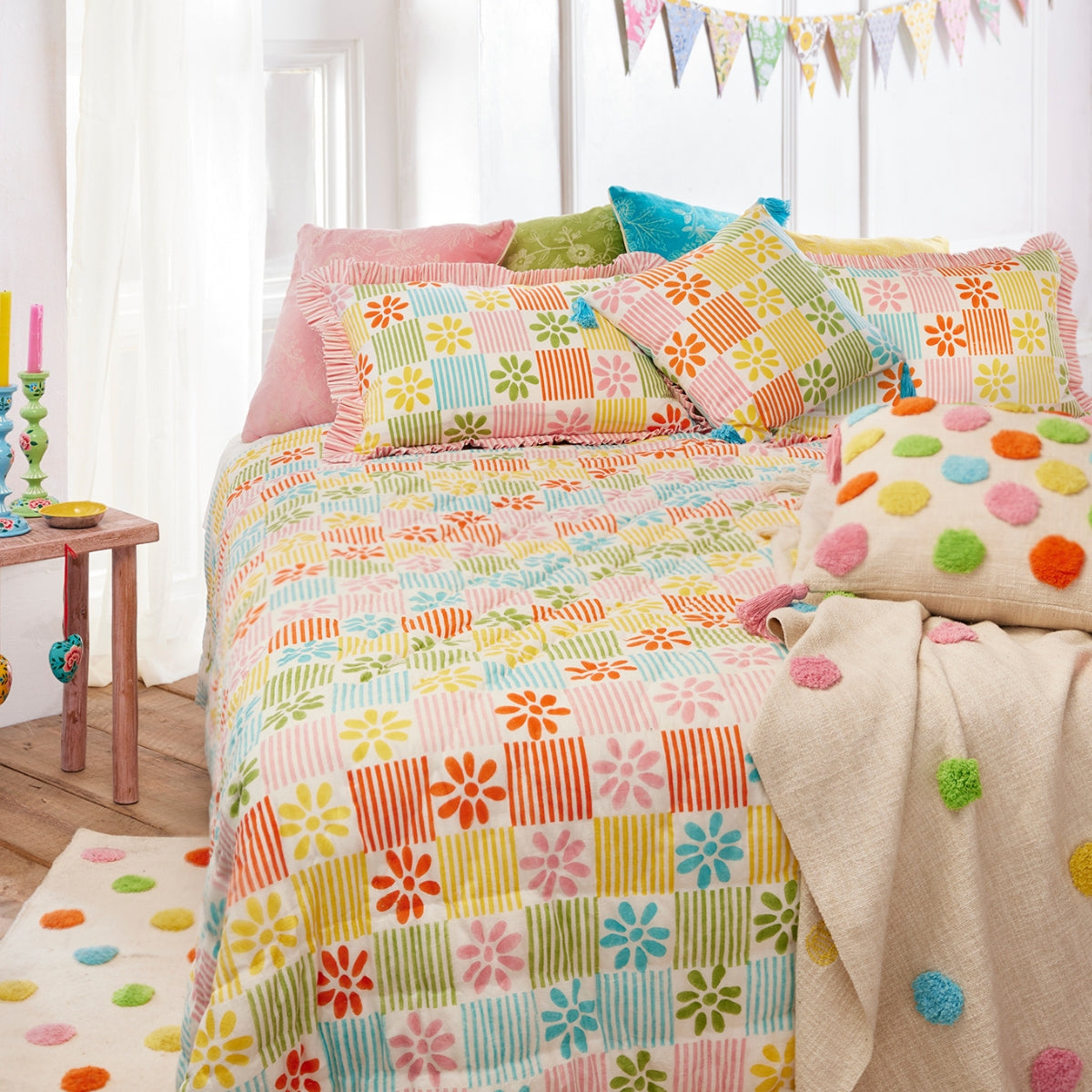 A colorful patchwork quilted bedding set styled on a bed in a bright bedroom. The set features a multicolor floral and striped pattern in pink, orange, yellow, green, and blue, with matching decorative pillows and a beige throw blanket with colorful pom-poms.