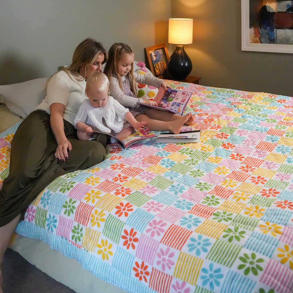 A woman, two children, and a baby sitting on a bed reading a book, showcasing a vibrant, multicolored retro-style floral quilted coverlet. The bedding features a patchwork design of pink, blue, yellow, green, and orange squares with floral and striped patterns.