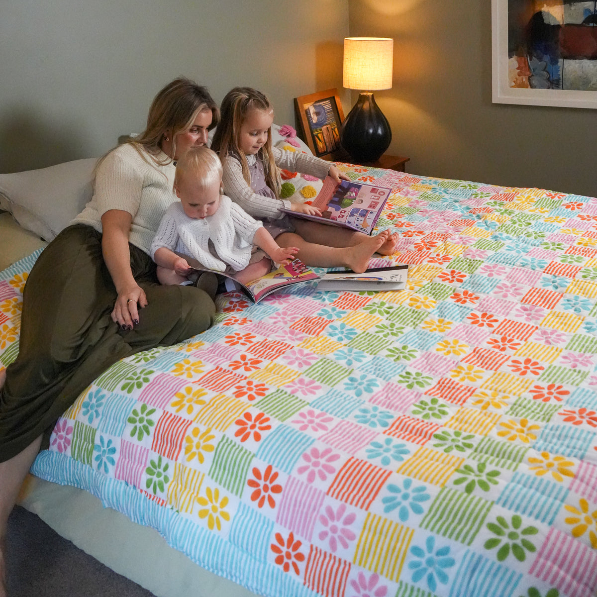 A woman, two children, and a baby sitting on a bed reading a book, showcasing a vibrant, multicolored retro-style floral quilted coverlet. The bedding features a patchwork design of pink, blue, yellow, green, and orange squares with floral and striped patterns.