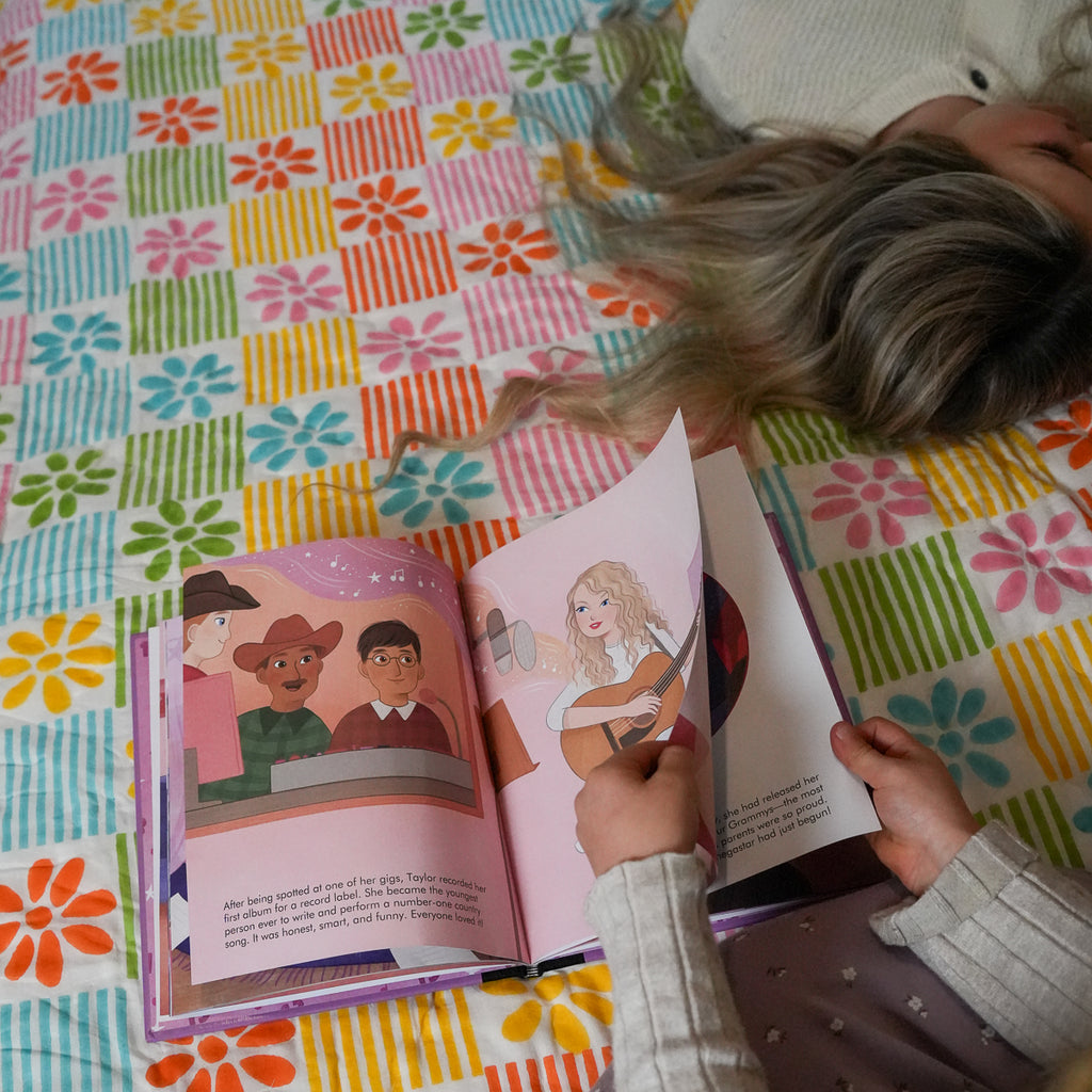A child's hands holding an open illustrated book on a colorful floral checkered blanket, with a person lying down nearby.