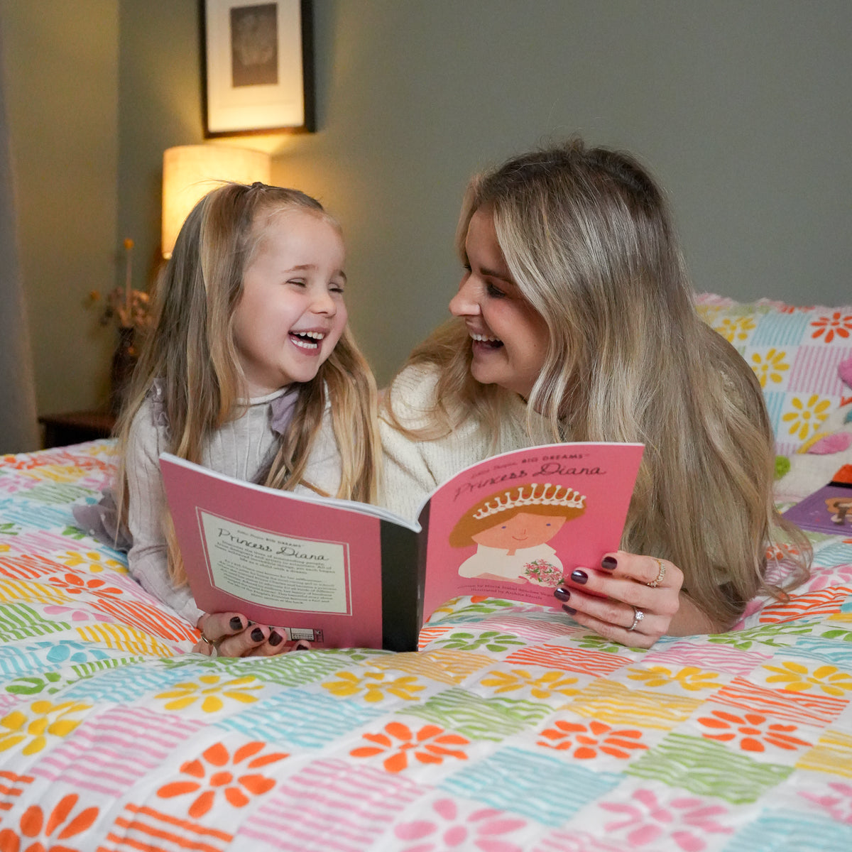 A woman and a young girl are lying on a bed reading a pink children's book together. The room has a colorful floral-patterned bedspread, and a lamp is visible in the background.