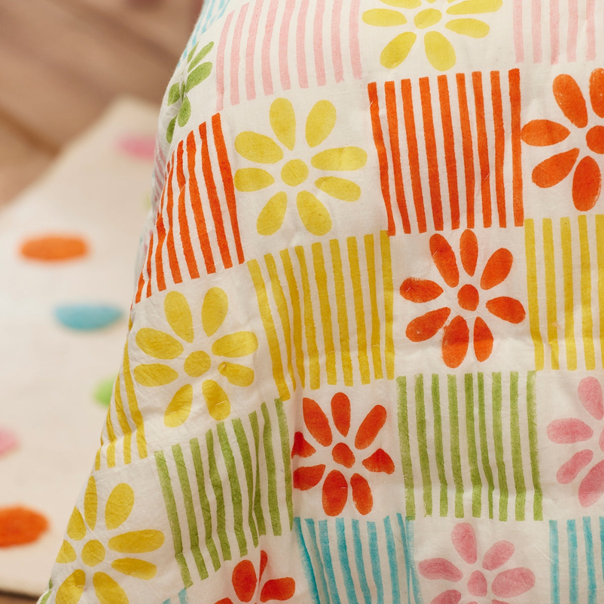 A close-up of a quilted blanket featuring a retro patchwork pattern of colorful flowers, stripes, and solid blocks in yellow, orange, green, pink, and blue on a white background.