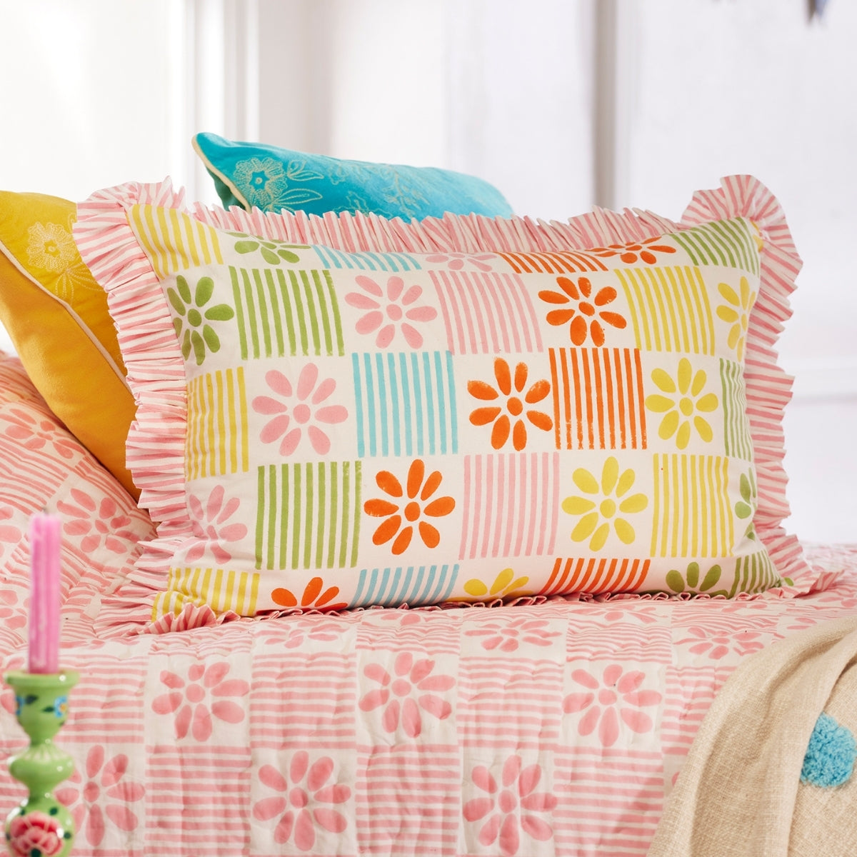 A decorative throw pillow with a colorful patchwork floral and striped pattern, styled on a bed with a pink floral quilt and other accent pillows.