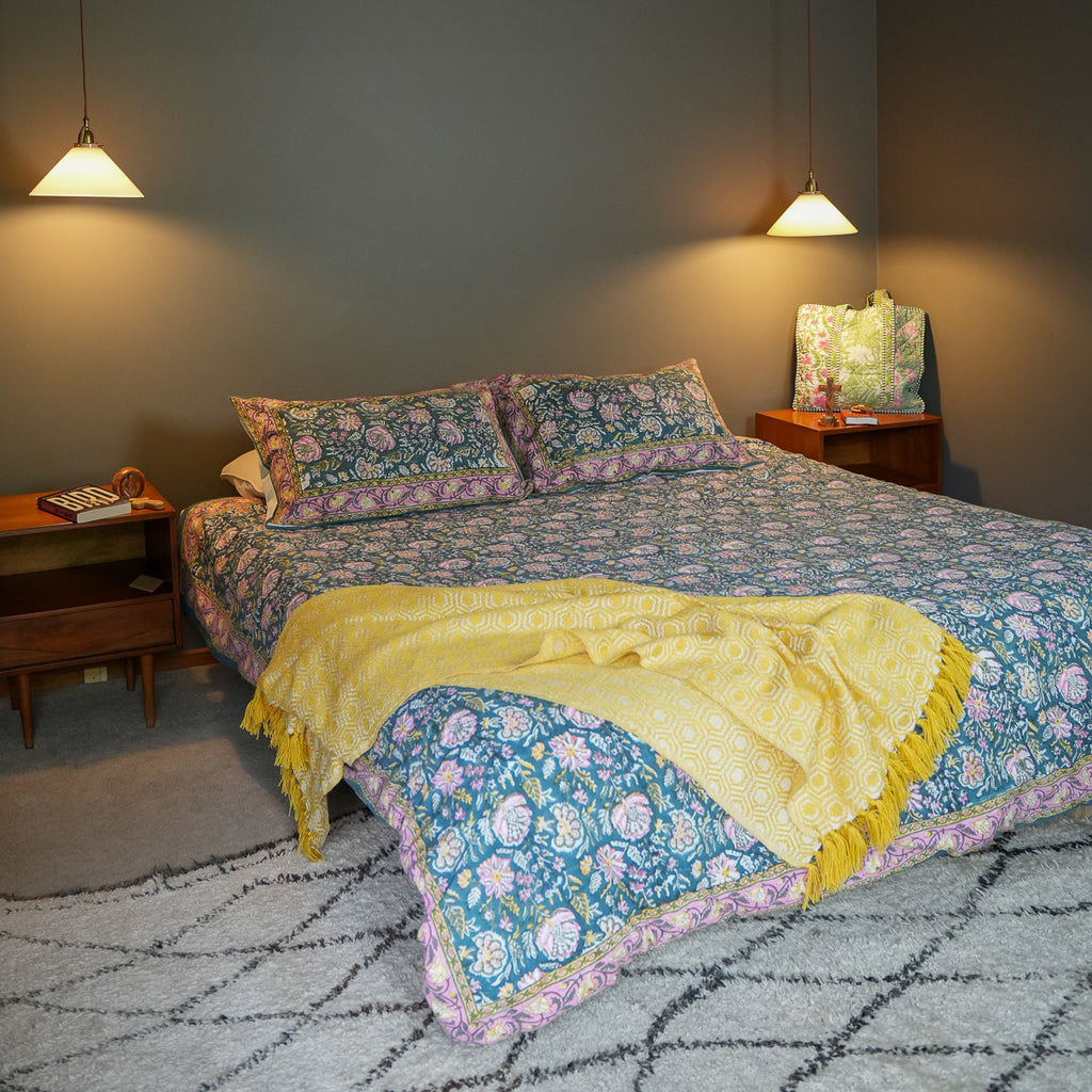 A bed dressed in a blue floral quilted coverlet with matching pillowcases, a yellow fringed throw blanket, and a patterned pillow, set in a styled bedroom with wooden nightstands, hanging lamps, and a gray wall.