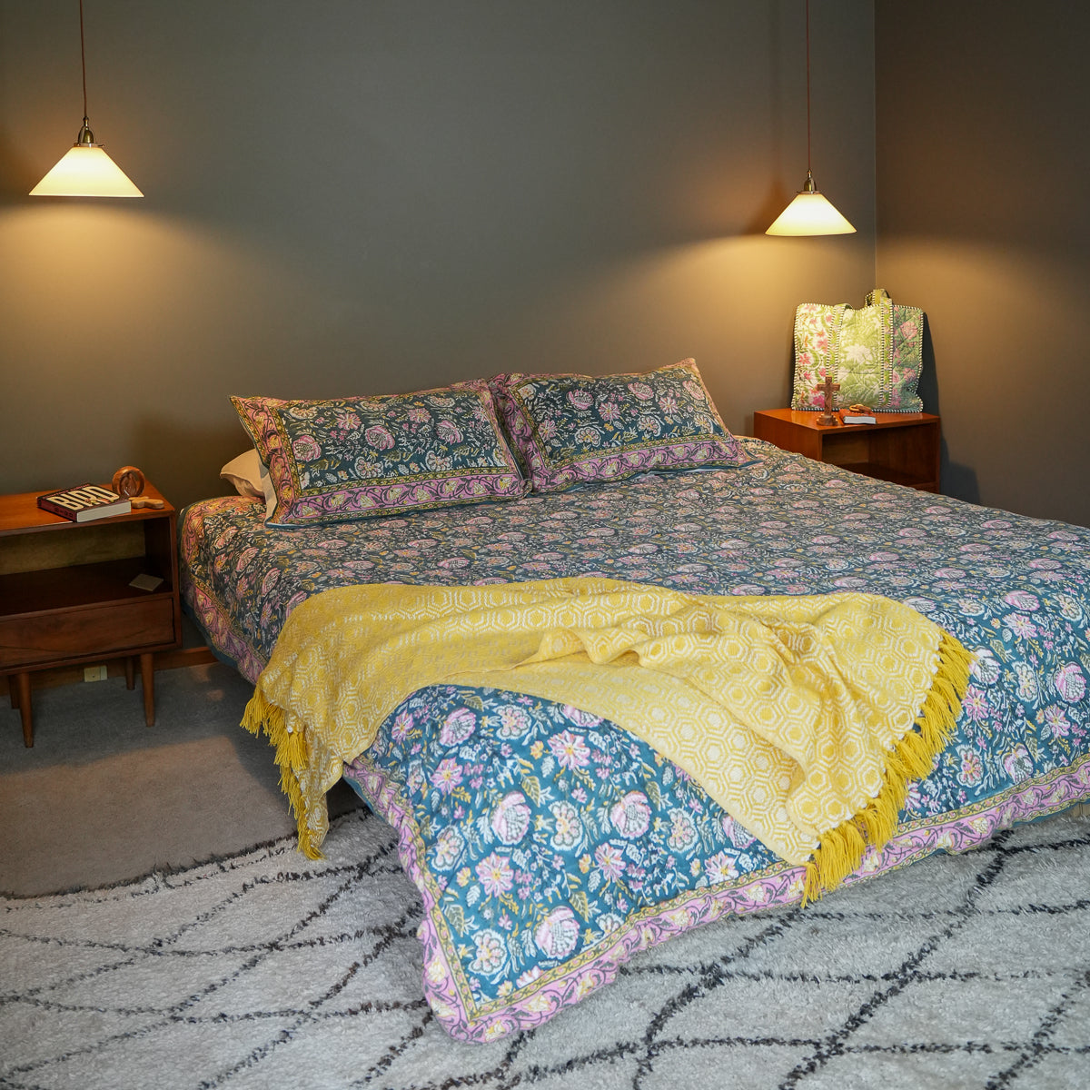 A bed dressed in a blue floral quilted coverlet with matching pillowcases, a yellow fringed throw blanket, and a patterned pillow, set in a styled bedroom with wooden nightstands, hanging lamps, and a gray wall.