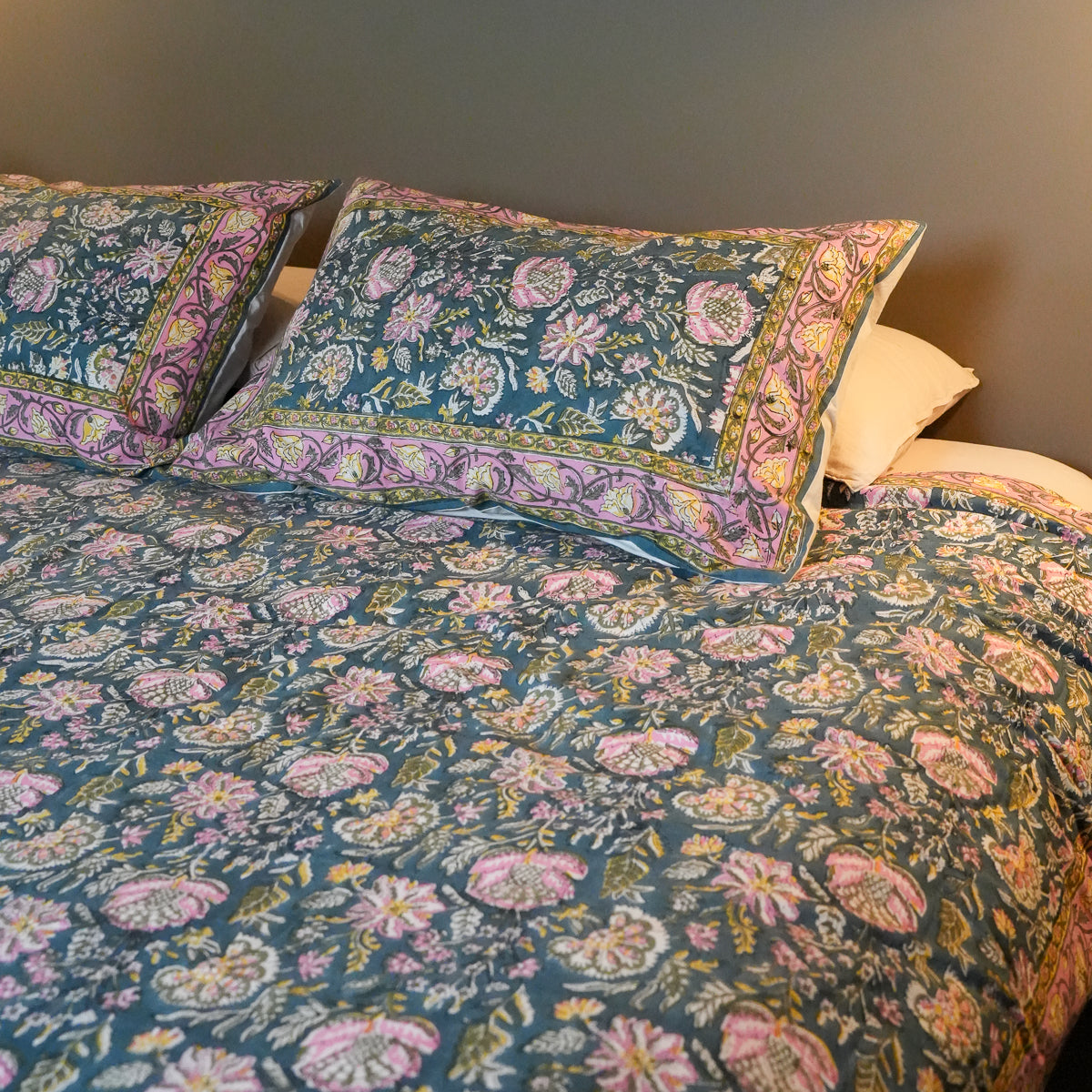 A bed dressed in a teal floral duvet cover and matching pillowcases with pink, purple, and gold ornamental borders, shown in a bedroom setting against a dark gray wall.