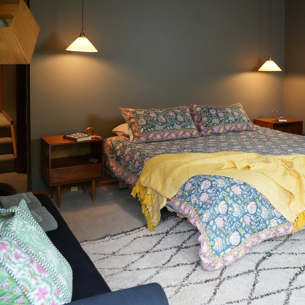 A cozy bedroom scene featuring a bed dressed in a blue and pink floral patterned quilt and matching pillow shams, with a bright yellow throw blanket draped across the foot. The room has a dark gray wall, wooden bedside tables, hanging pendant lights, and a patterned rug.