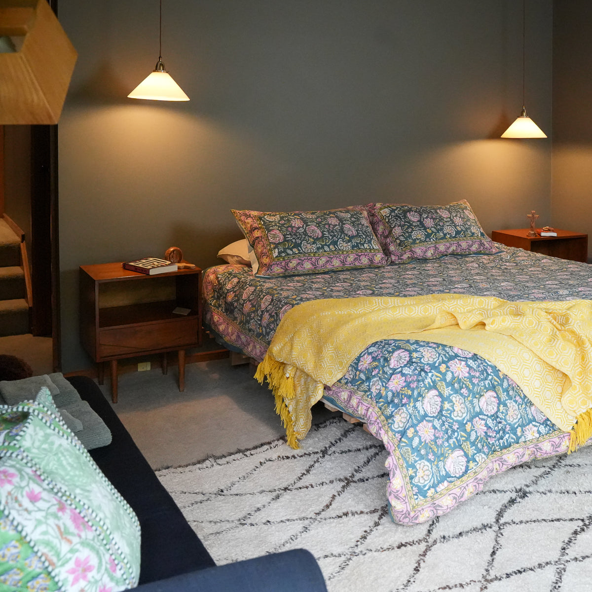A cozy bedroom scene featuring a bed dressed in a blue and pink floral patterned quilt and matching pillow shams, with a bright yellow throw blanket draped across the foot. The room has a dark gray wall, wooden bedside tables, hanging pendant lights, and a patterned rug.