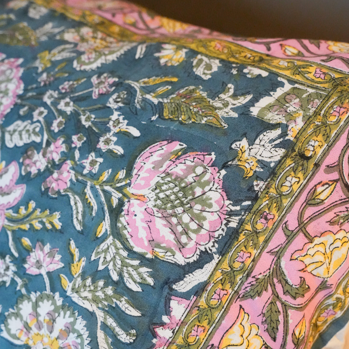 A close-up of a patterned textile or cushion cover featuring a teal blue background with pink, yellow, and white floral motifs and a decorative border.