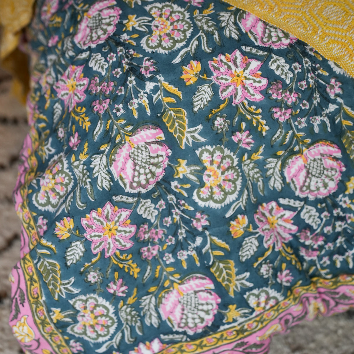 A close-up of a floral printed textile, likely a skirt or dupatta, featuring a teal blue-green base with pink, yellow, and white block-printed flowers and leaves, finished with a pink and yellow border.