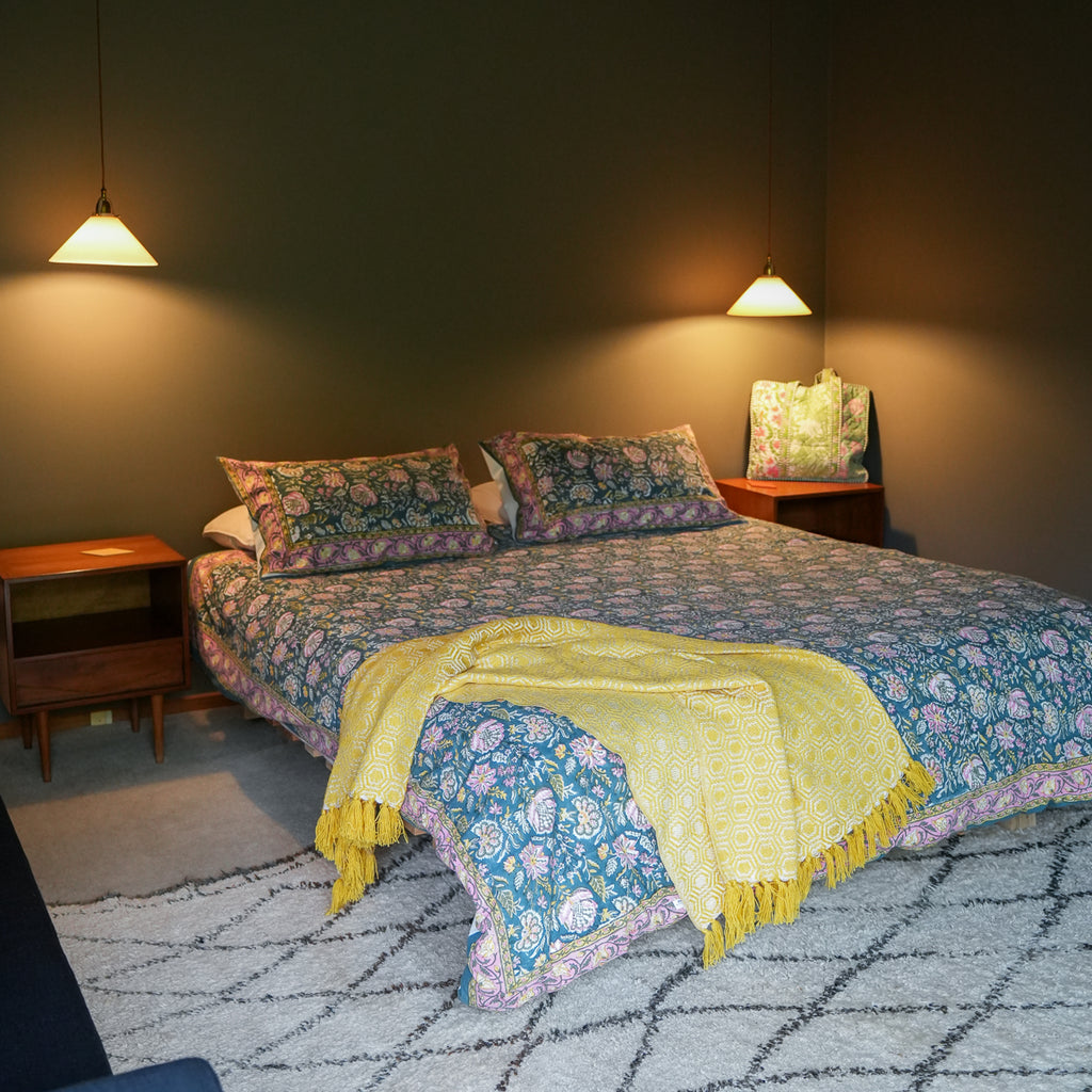 A bed dressed in a blue floral-patterned duvet cover and matching pillows, with a bright yellow fringed throw blanket draped across the foot, set in a styled bedroom with wooden nightstands, pendant lights, and a patterned rug.