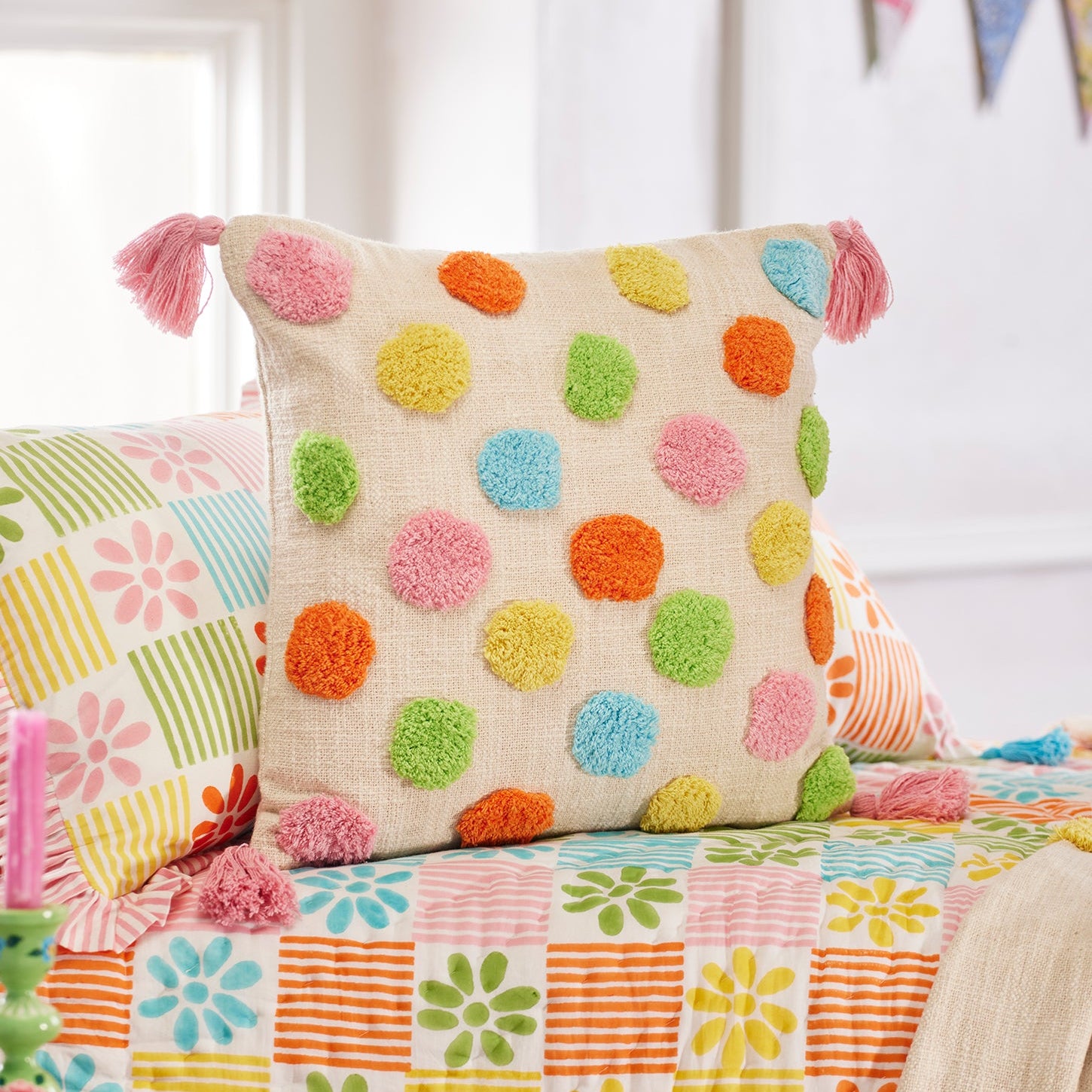 A decorative throw pillow with colorful textured pom-pom dots on a cream linen background, styled on a bed with a matching floral quilt and other pillows in a bright bedroom setting.