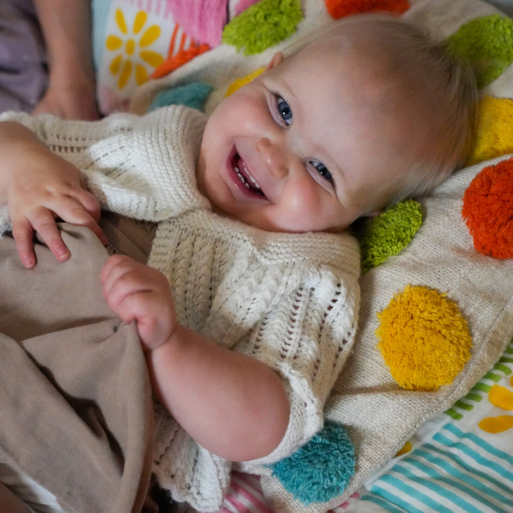 A smiling baby lying on a colorful knit blanket with pom-poms, wearing a cream-colored knit top and brown pants.