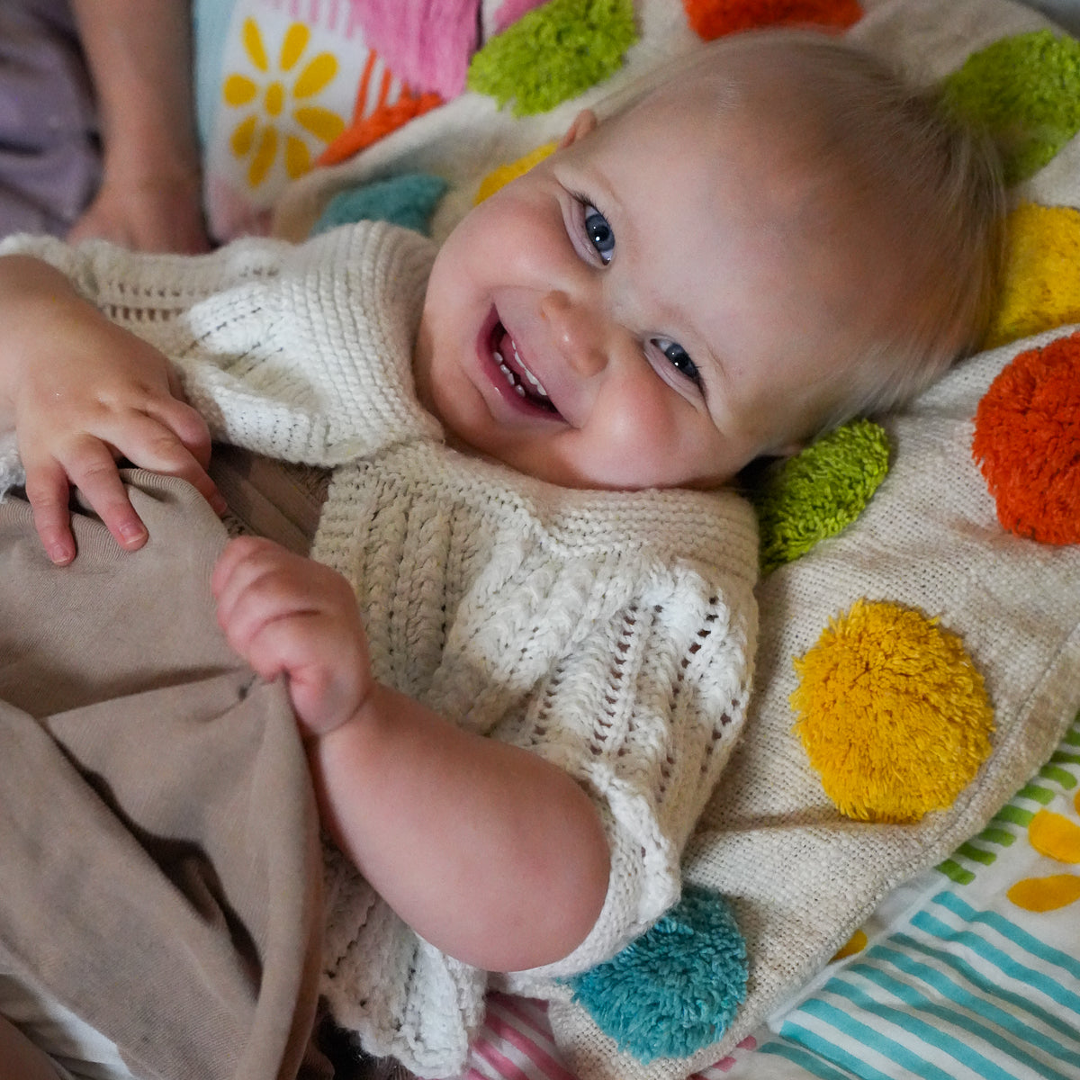 A smiling baby lying on a colorful knit blanket with pom-poms, wearing a cream-colored knit top and brown pants.