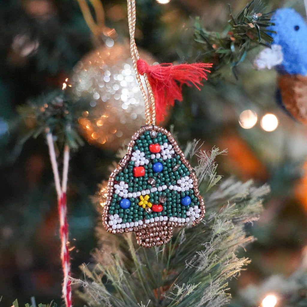 A Christmas tree ornament made of green, white, red, blue, and yellow beads hangs from a gold string on a decorated Christmas tree branch.