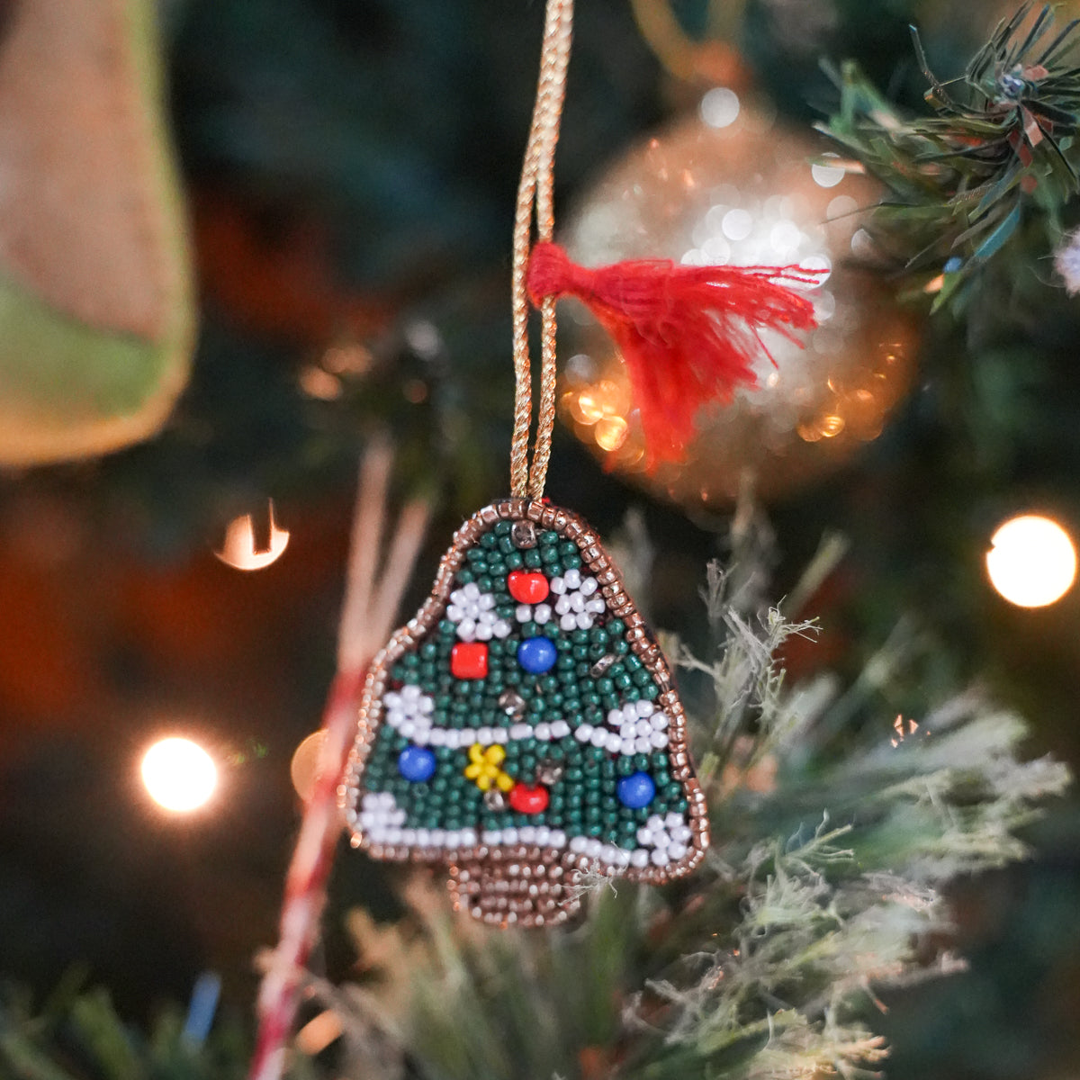 A beaded Christmas tree ornament hanging on a decorated Christmas tree branch. The ornament is made of green, white, and blue beads with small red and yellow accents, hanging from a gold cord with a red tassel.