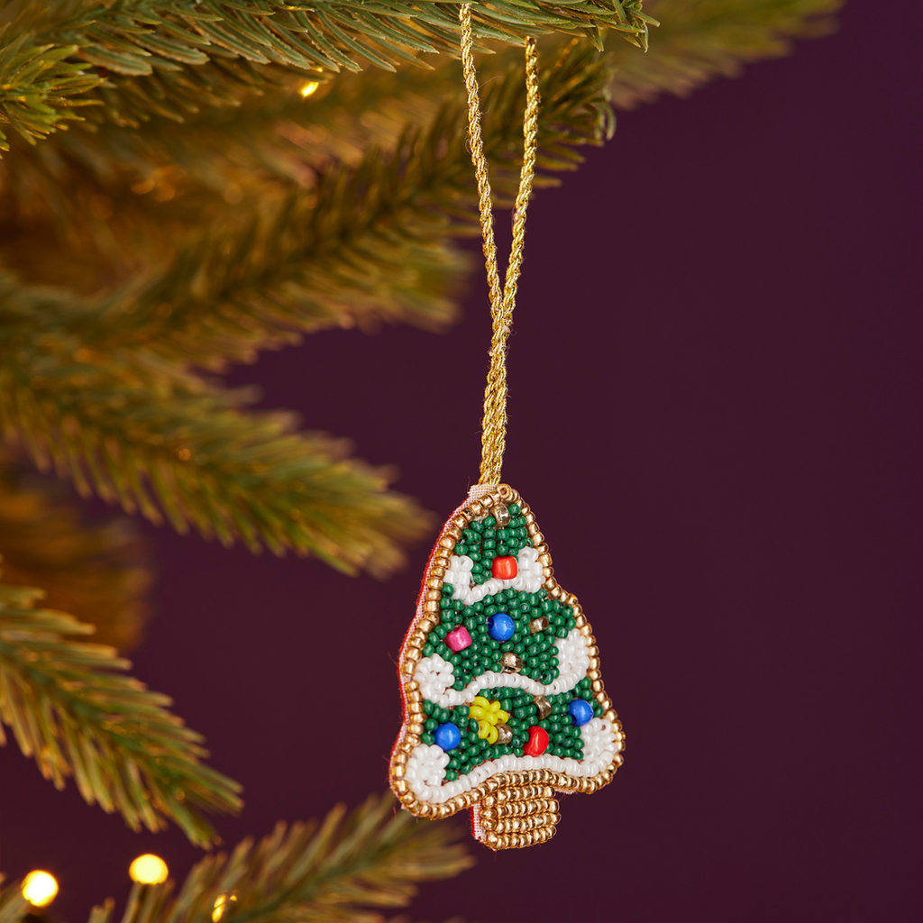 A hand-beaded Christmas tree ornament hanging from a gold string on a Christmas tree branch, with a dark purple background and warm bokeh lights.