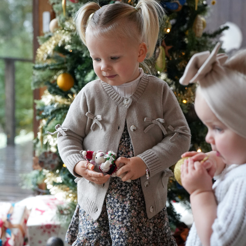 A toddler girl in a beige knit cardigan with bow details and a floral dress stands in front of a decorated Christmas tree, holding small plush toys. Another child is partially visible on the right in a cozy indoor holiday setting.
