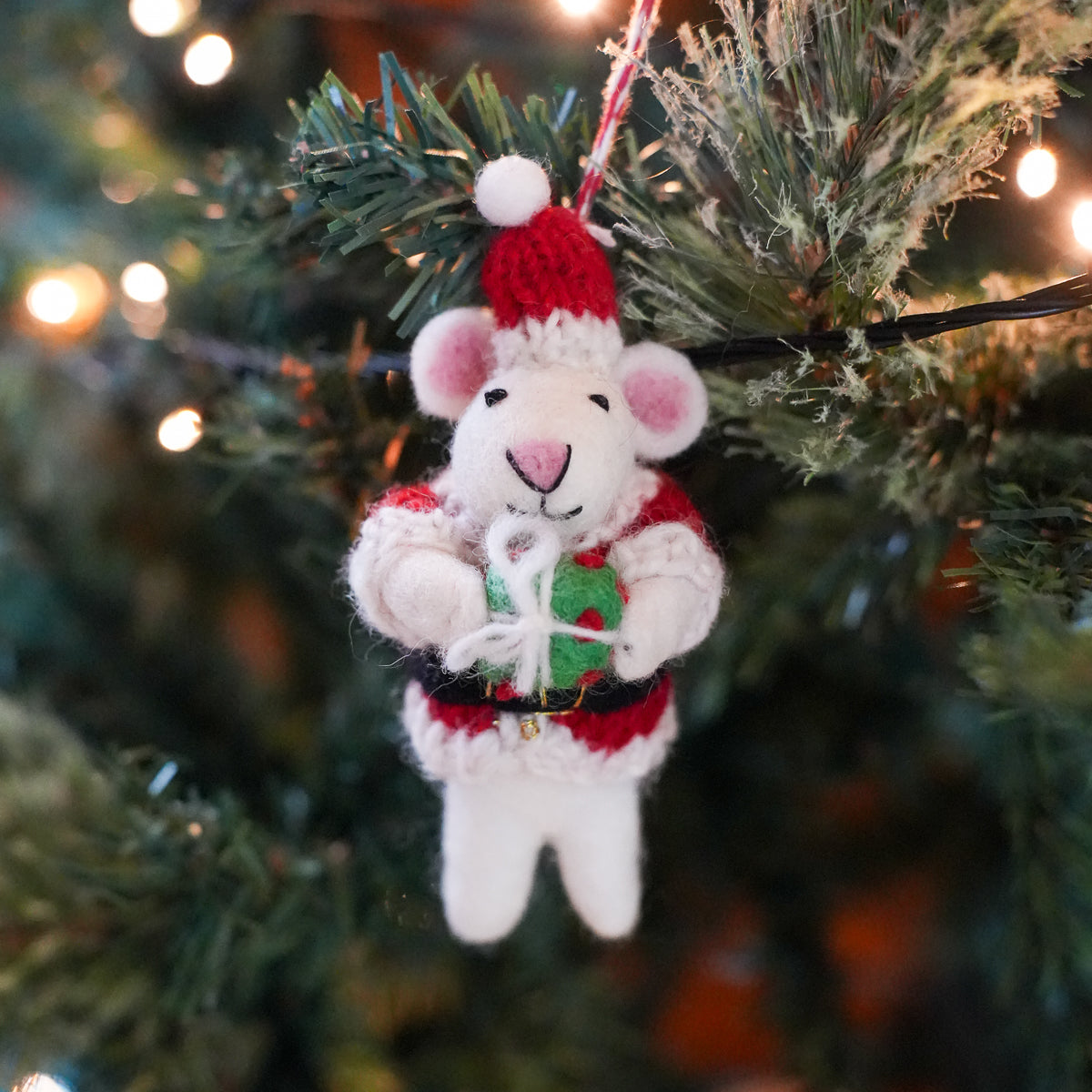 A handmade felt Christmas ornament shaped like a white mouse dressed in a red Santa hat and Santa suit, holding a green gift box with a white bow, hanging on a decorated Christmas tree branch with warm lights in the background.