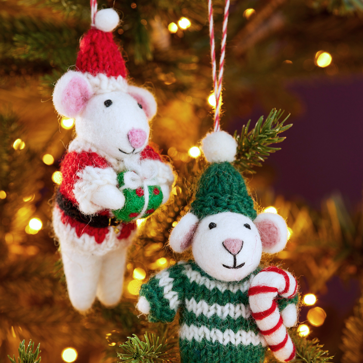 Two knitted mouse Christmas ornaments hanging on a blurred Christmas tree with warm bokeh lights. One mouse wears a red Santa hat and red-and-green outfit holding a wrapped gift, and the other wears a green knit hat and green-and-white striped sweater holding a red-and-white candy cane.