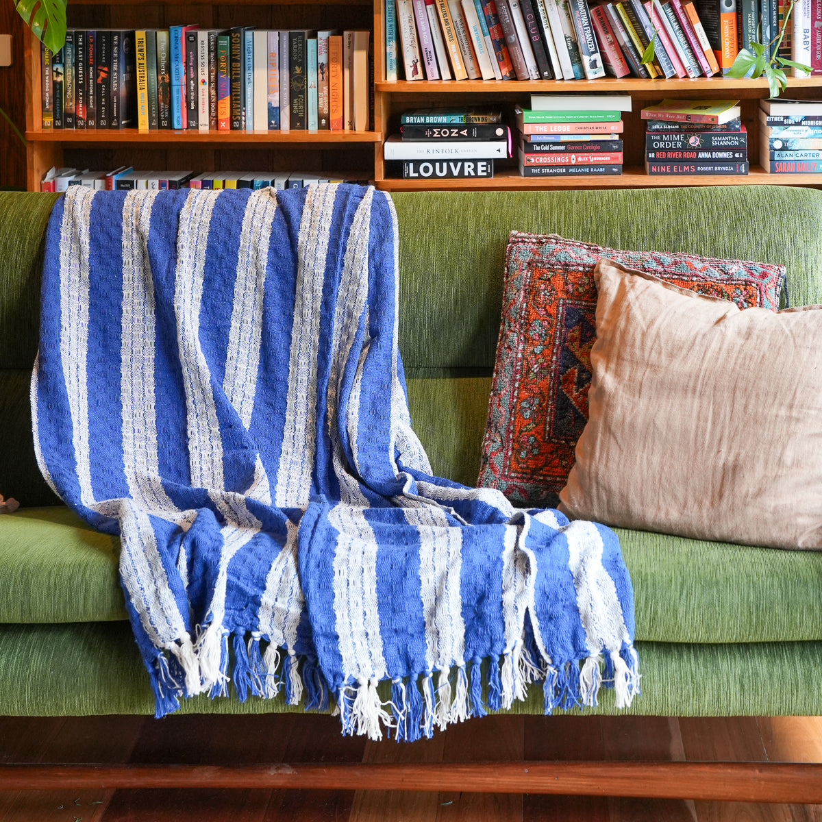A blue and white striped fringed throw blanket is draped over a green sofa in a cozy living room setting, with patterned and beige pillows and a wooden bookshelf in the background.