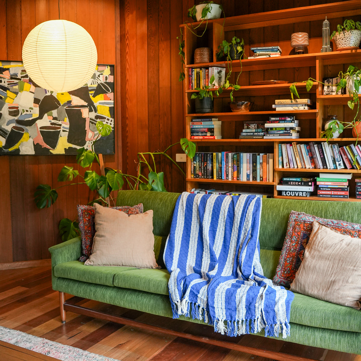 A green sofa in a mid-century modern living room, styled with a blue and white striped throw blanket, beige pillows, and a wooden bookshelf filled with books and plants in the background.