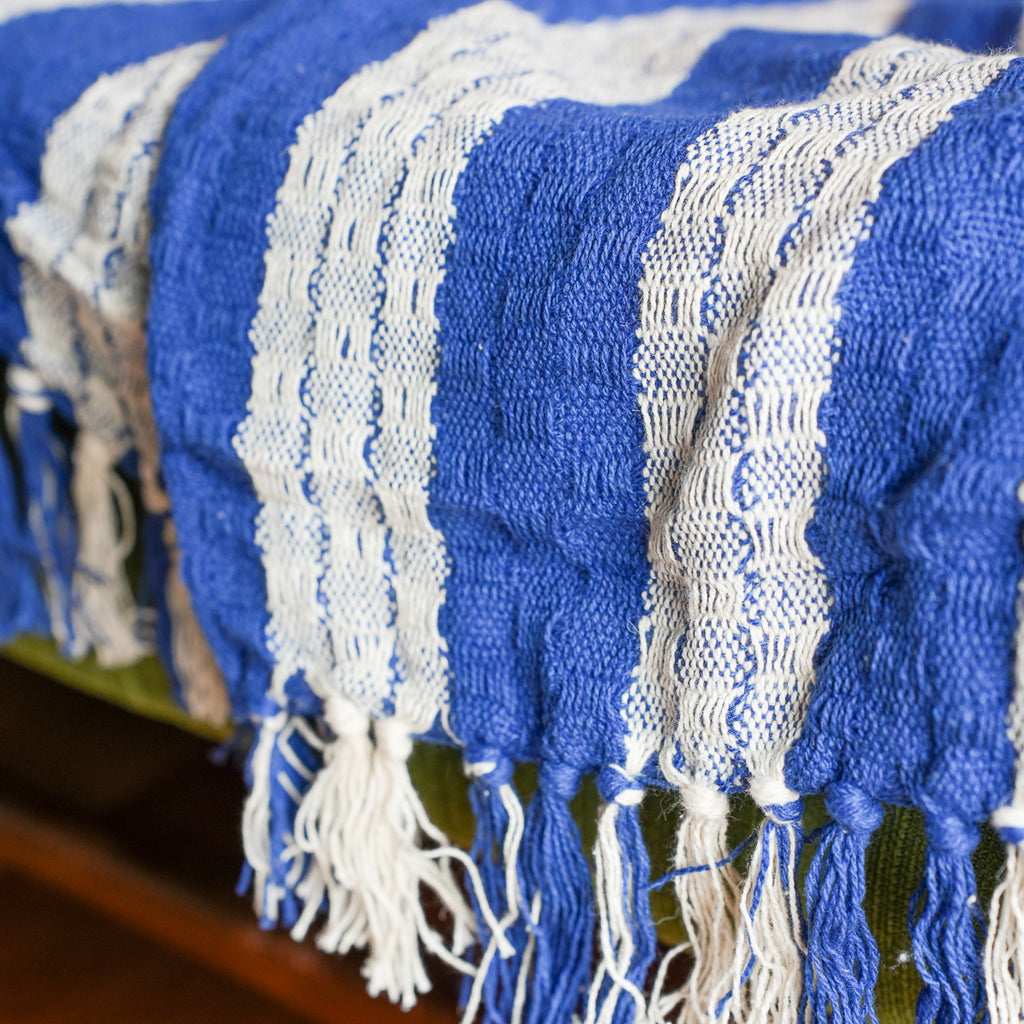 A close-up of a woven throw blanket with bold blue and white stripes and fringed tassels, draped over a surface.