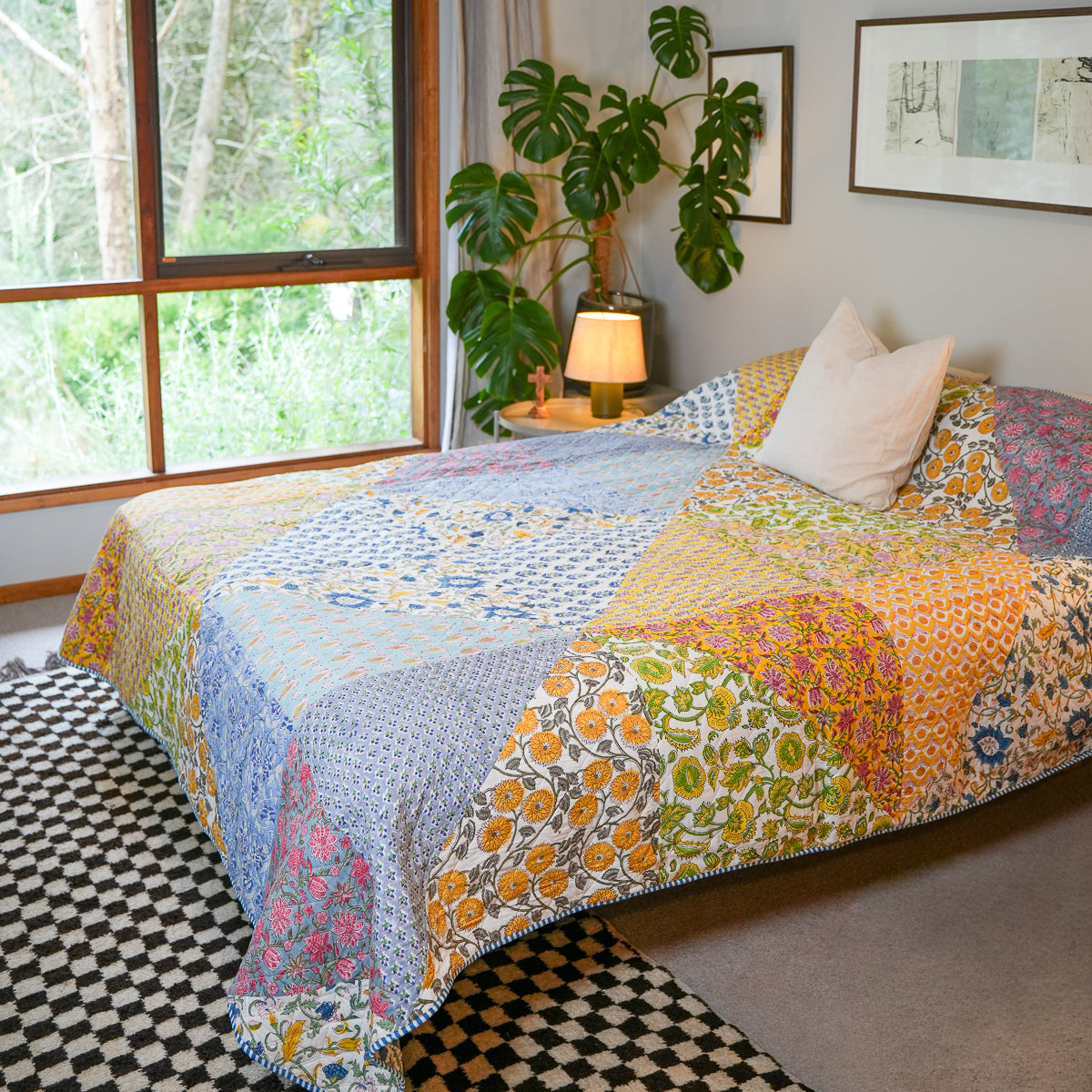 A colorful patchwork quilted bedspread with various floral and geometric patterns in blue, yellow, orange, pink, and green, styled in a bright bedroom next to a window and a potted plant.