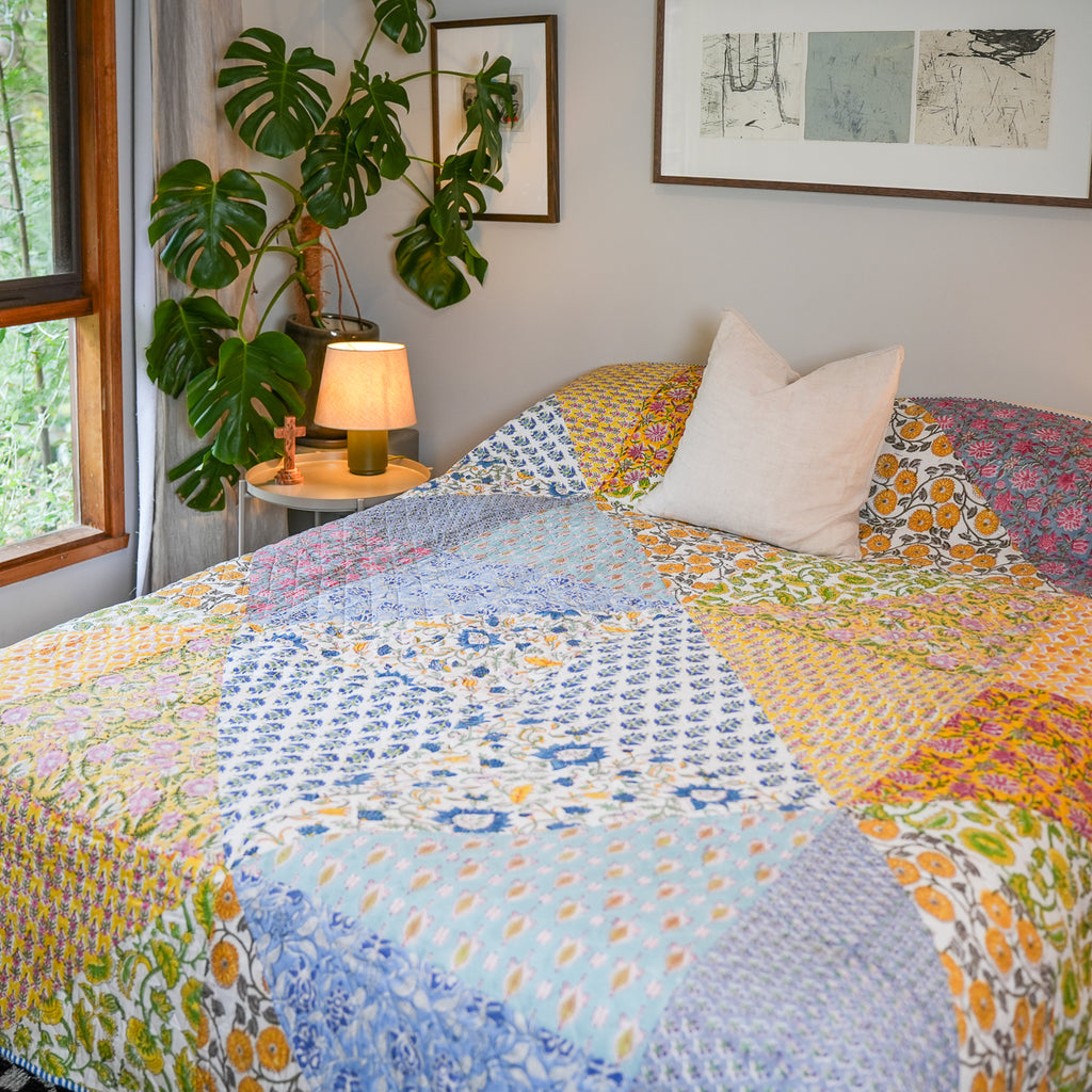 A colorful patchwork quilted bedspread with various floral and geometric patterns in blue, yellow, pink, and green, styled on a bed in a bright bedroom with a white pillow, a bedside lamp, and a potted monstera plant.