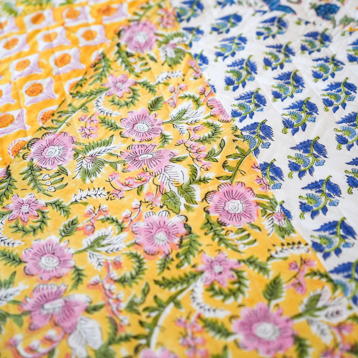 A close-up of a colorful printed fabric with three distinct patterns: a bright yellow floral section with pink flowers and green leaves, a white section with blue floral motifs, and a yellow section with orange circular patterns.