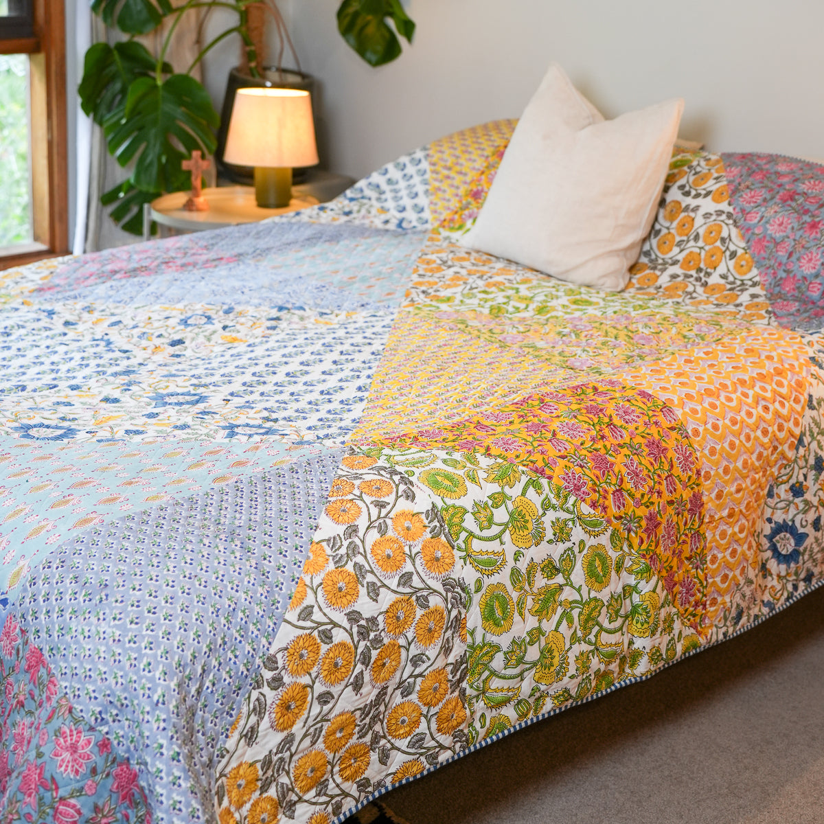 A colorful patchwork quilted bedspread with various floral and geometric patterns in orange, yellow, blue, pink, and green, styled on a bed with a white pillow in a cozy bedroom setting.